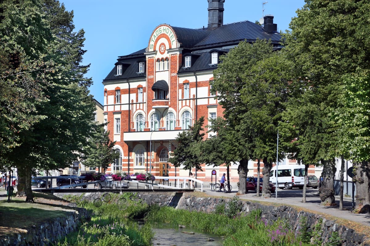 En stor, röd tegelsbyggnad med vita detaljer och svarta tak dominerar bilden. Byggnaden, märkt med texten ”STADSHOTEL” över entrén, omges av lummiga gröna träd. En person med cykel syns på en gångväg vid sidan av en liten, stenbelagd kanal nedanför b