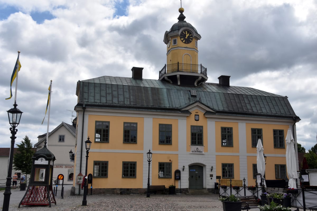 En gul byggnad med ett klocktorn och ett mörkgrönt tak under en molnig himmel. Framför byggnaden finns en kullerstenstorg med gatlyktor, flaggstänger med gula och blåa flaggor, samt en svart "Rikstelefon"-kiosk. Några vitklädda parasoller syns till h