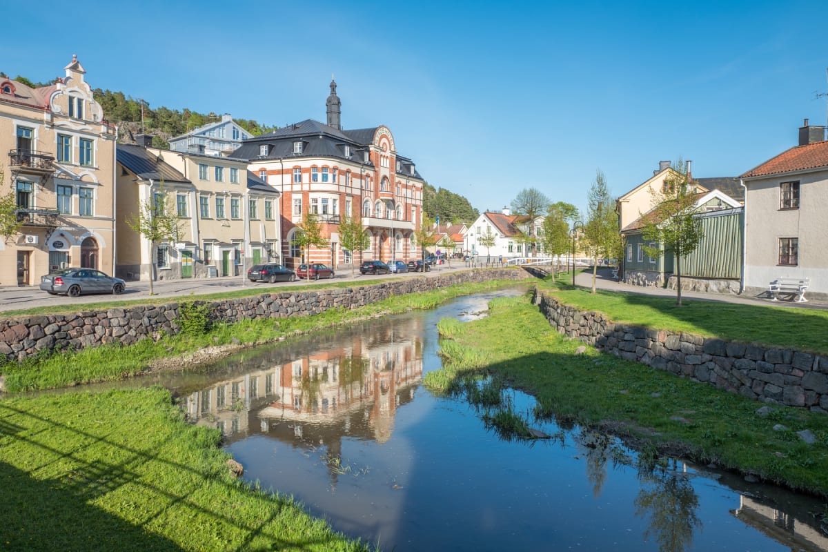 En gata längs en flod eller å med hus i bakgrunden. Vissa hus har äldre arkitektur med detaljer som takspiror och burspråk. Gräs och träd finns längs vattnet och gatan. Bilar är parkerade längs vägkanten. Himlen är klarblå.