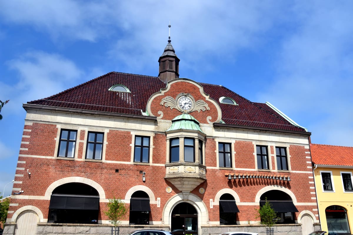 En stor, röd tegelbyggnad med ett brunt tak syns under en blå himmel med vita moln. Byggnaden har en tidstypisk design med en centralt placerad klocka, en kupolformad del med ett burspråk och en spira på taket. Fönster med svarta ramar framträder tyd