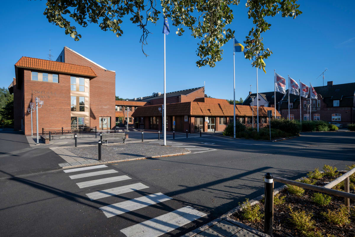 En fasad av rött tegel som tillhör Tomelilla kommun är synlig under en klarblå himmel. Framför fasaden finns ett övergångsställe, träd med gröna löv och flaggor, inklusive den svenska och EU-flaggan.
