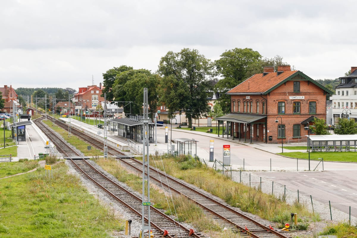 En översiktsbild av en järnvägsstation i Tomelilla under dagtid med tågspår i förgrunden, perronger, och en tegelbyggnad med texten "TOMELILLA" i bakgrunden. Ett gräsbevuxet område fyller delar av förgrunden.