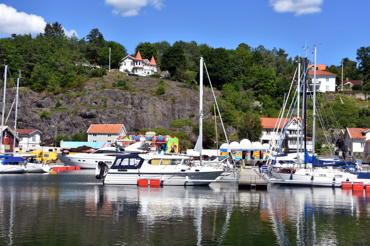 En bild som visar en småbåtshamn under en solig dag. I förgrunden ligger flera båtar förtöjda vid bryggor. Bakom hamnen reser sig en klippig sluttning med bebyggelse och lummig grönska. Himmelen är blå med några vita moln.