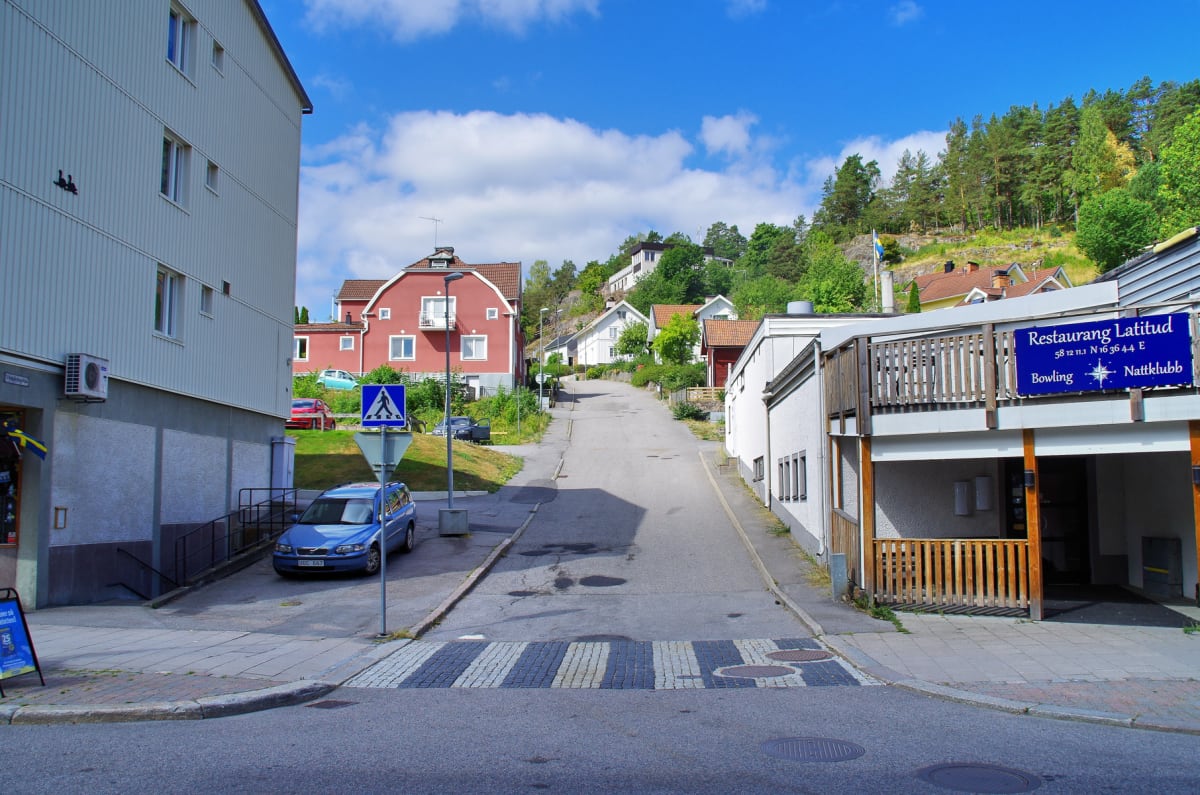 En gata framför en byggnad med en blå bil parkerad vid trottoaren. Till höger syns en byggnad med en skylt som anger "Restaurang Latitud". Längre bort syns en backe med hus och träd. En fotgängarskylt och ett övergångsställe syns i förgrunden.