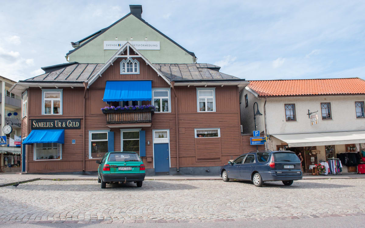 En gata med gatsten framför flera byggnader, där en av byggnaderna har texten "SAMELIUS UR & GULD" och blå markiser. Två bilar, en grön och en mörkblå, är parkerade på gatstenen. Ovanför syns en ljusblå himmel med några moln.
