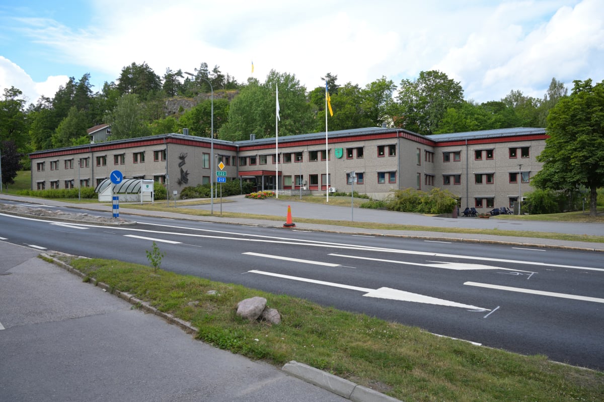 En tvåvåningsbyggnad med grå fasad och röda detaljer står intill en väg med vita pilar och markeringar. Grönskande träd och buskar omger byggnaden och en kulle syns i bakgrunden. Två flaggstänger med flaggor är placerade framför byggnaden.