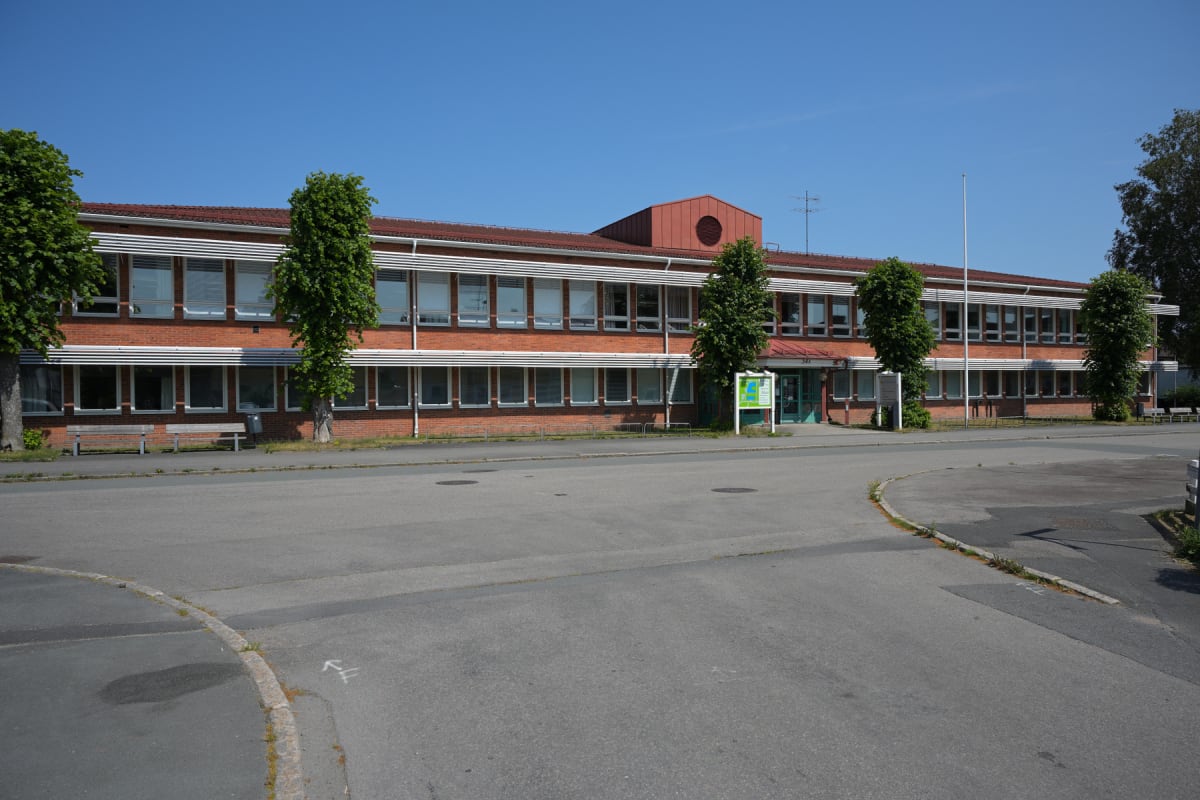 En stor, röd tegelbyggnad med många fönster och vita persienner under en klarblå himmel. Framför byggnaden finns en asfalterad väg med trottoarer, några träd med gröna löv, bänkar och en informationsskylt vid en ingång.