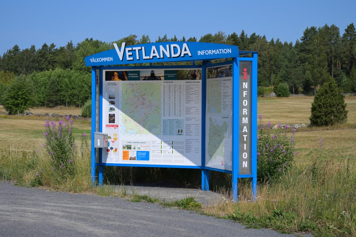 En blå informationsskylt med texten "VÄLKOMMEN VETLANDA INFORMATION" står vid kanten av en grusväg. Skylten innehåller kartor och information om Vetlanda. I bakgrunden syns ett grönt skogsbryn och en gräsbevuxen åker med vissna partier. Vid sidan av