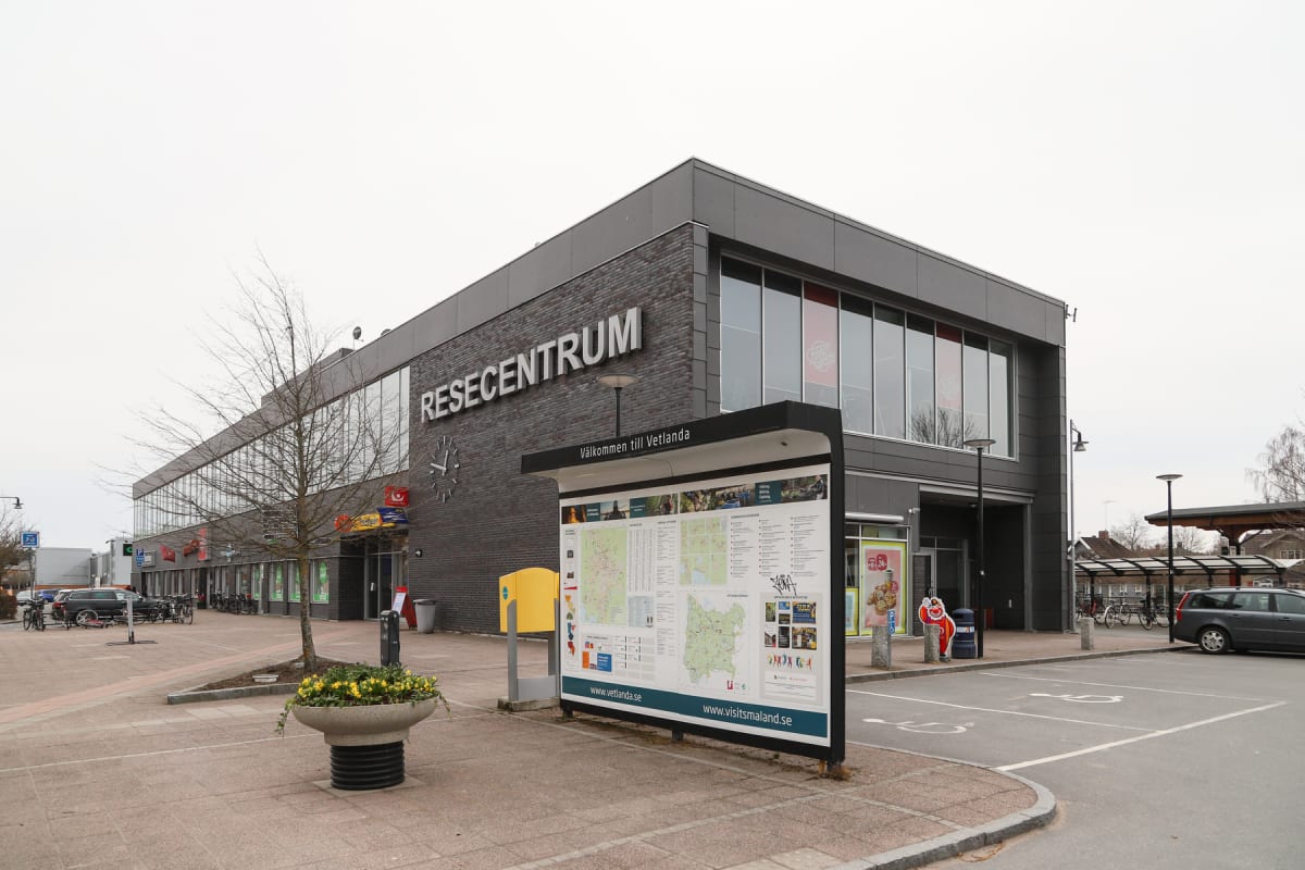 En modern byggnad med texten "RESECENTRUM" på fasaden. Framför byggnaden finns ett "Välkommen till Vetlanda" skylt, cyklar parkerade, en stubbe med gula blommor och en bil parkerad. En kal träd står i förgrunden till vänster om byggnaden.