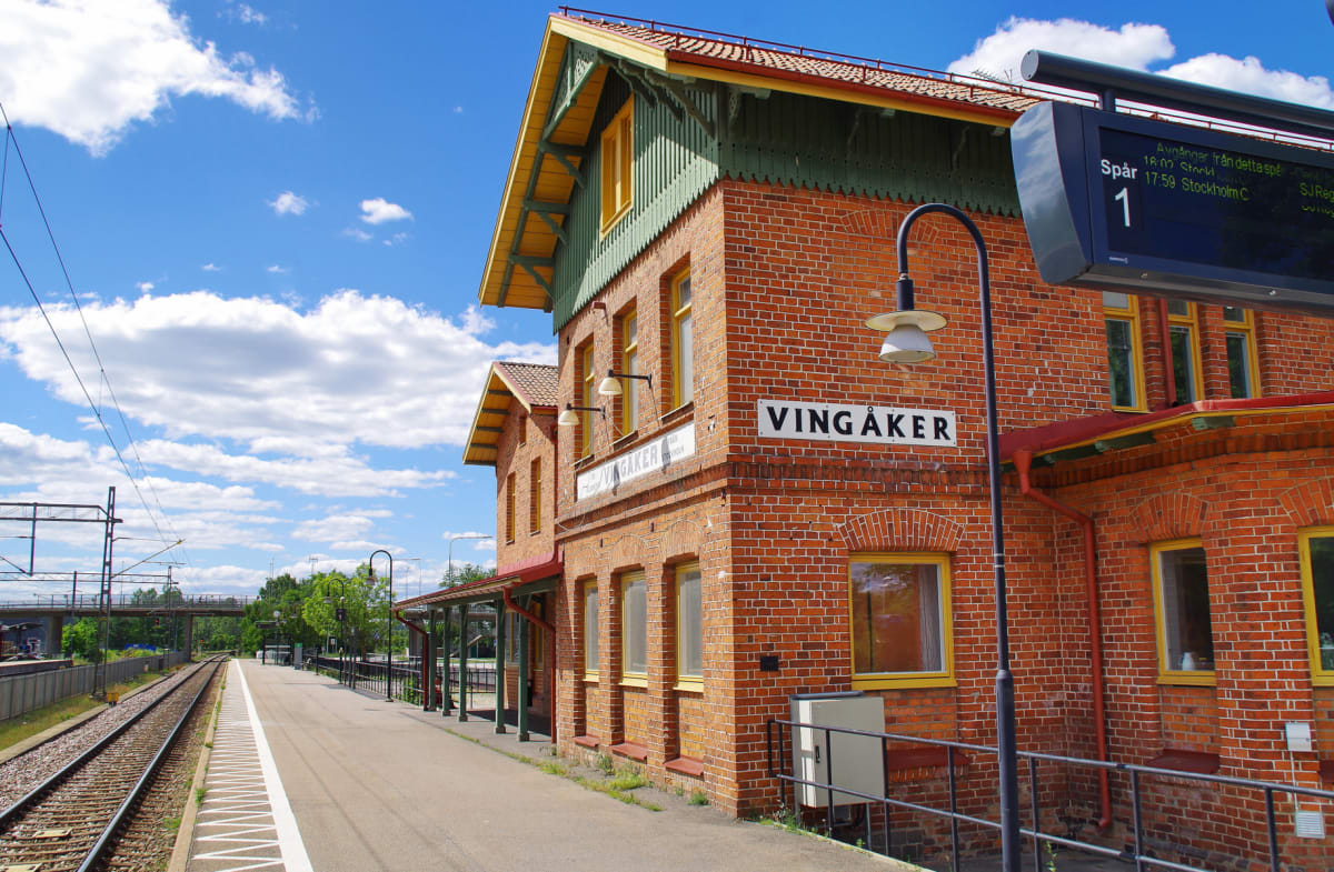 En bild av en järnvägsstation i Vingåker en solig dag. Ett rött tegelhus med gula fönster och en grön träfasad syns intill spåren. Till vänster löper järnvägsspår och en perrong, och i bakgrunden syns molnig himmel.