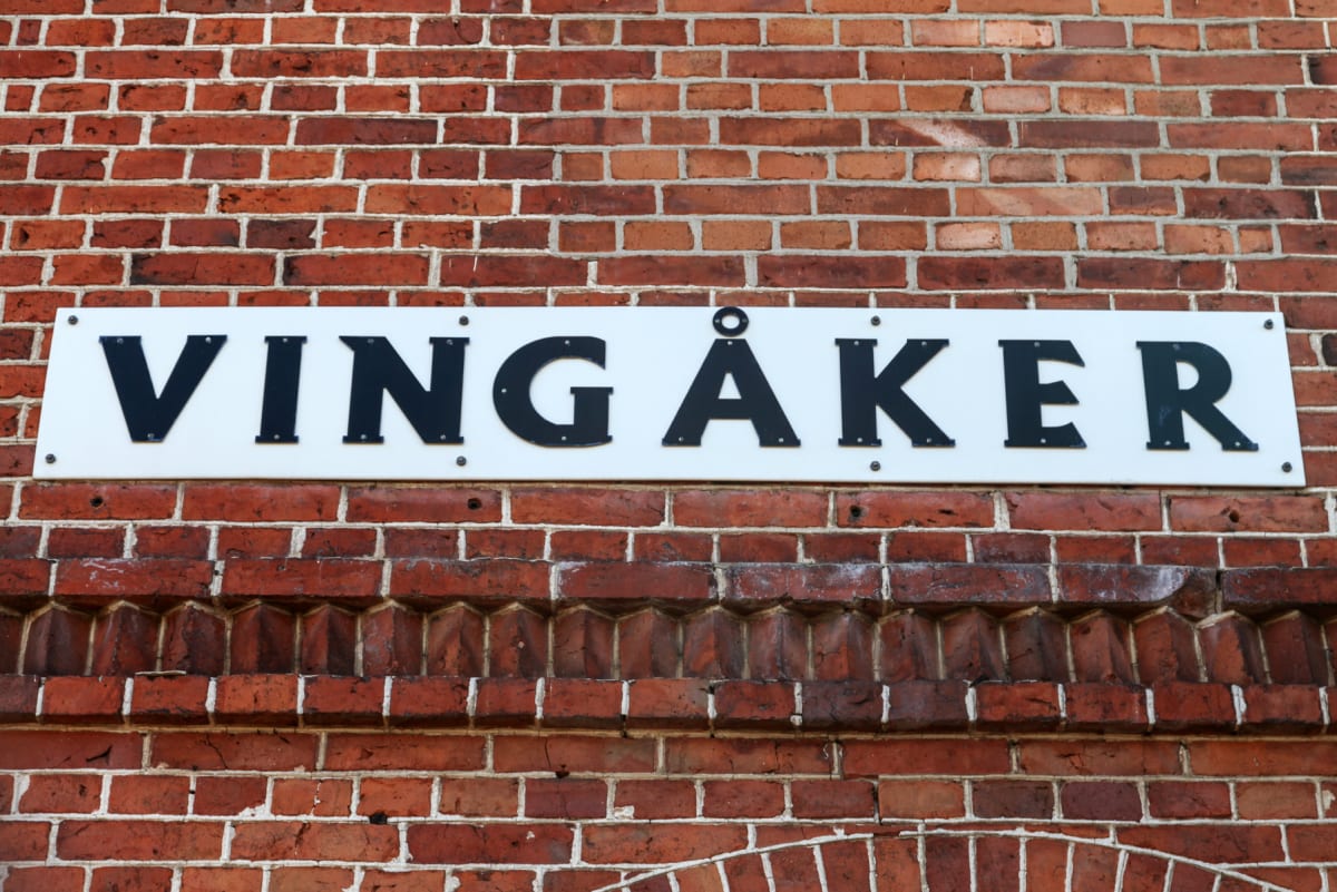 En vit skylt med de svarta bokstäverna ”VINGÅKER” monterad på en röd tegelvägg. Tegelväggen har detaljer i form av utskjutande stenar i en pyramidformad mönster. Inga människor eller fordon syns.