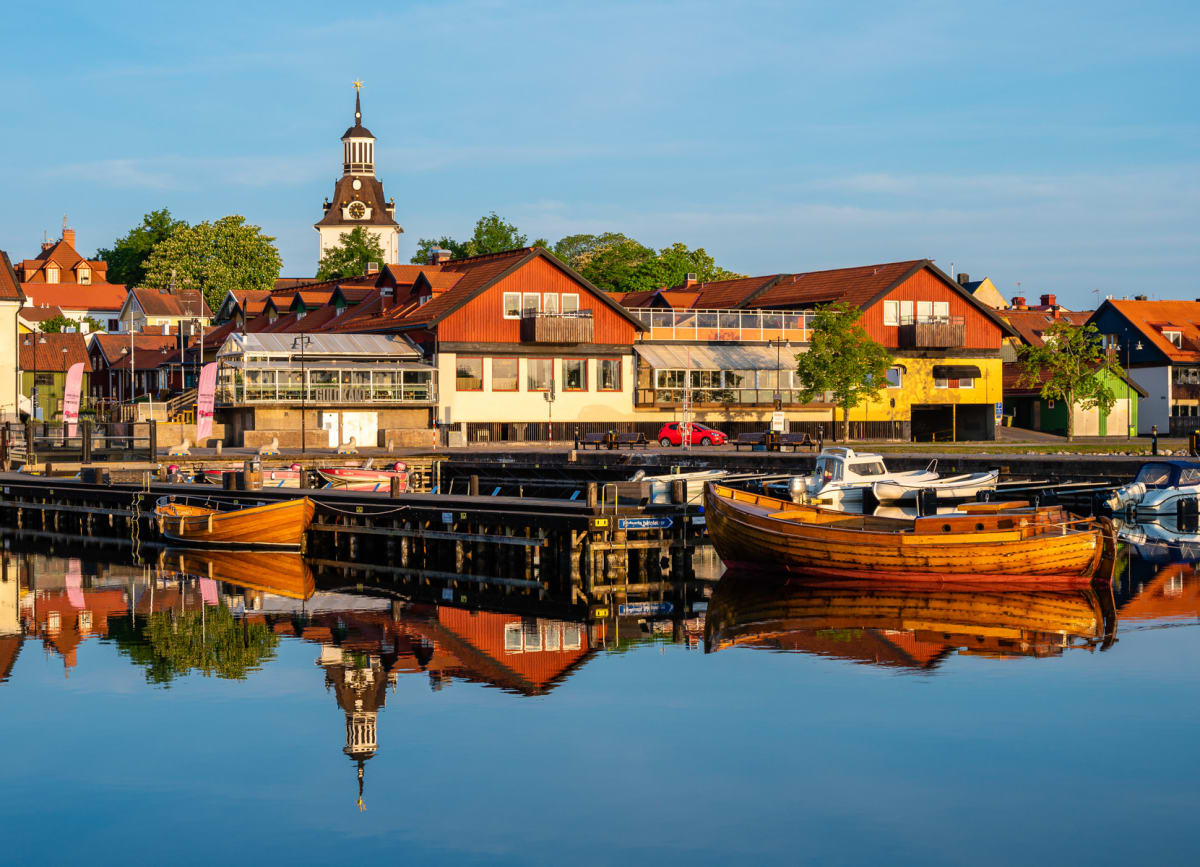 En bild visar en småbåtshamn med flera träbåtar och motorbåtar förtöja vid bryggor. I bakgrunden syns färgglada byggnader, inklusive ett klocktorn med ett spetsigt tak som reser sig över hustaken. Vattnet reflekterar båtarna, byggnaderna och skyn.