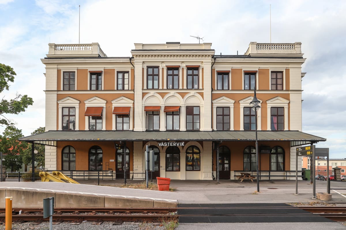 En stor, ljusbrun och vit byggnad med ett plåtat tak och texten "VÄSTERVIK" ovanför ingångarna. På taket syns en flaggstång utan flagga och en antenn. Framför byggnaden finns en perrong med räls, en röd papperskorg, en gatlykta och en bänk.