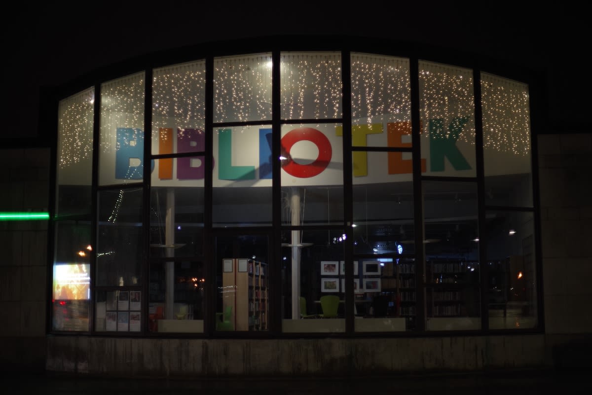 En fasadbild på en byggnad med stora fönsterpartier i mörker. Ovanför fönstren hänger ljusslingor och bakom glaset syns stora bokstäver som bildar ordet "BIBLIOTEK". Inuti byggnaden syns bokhyllor och inredning.