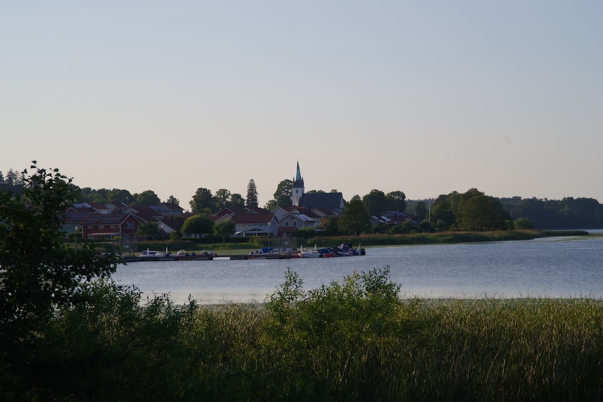 En översiktsbild av en sjö med ett samhälle längs strandlinjen i bakgrunden. Ett antal hus med röda tak syns, och i mitten av samhället reser sig en kyrka med en spetsig spir. Längs sjökanten ligger en brygga med båtar förtöjda. I förgrunden syns grä