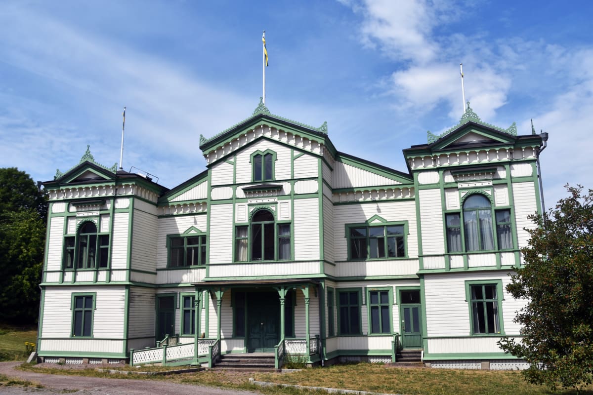 En stor vit byggnad med grönmålade detaljer står under en klarblå himmel med spridda moln. Byggnaden har flera fönster och en dörr på framsidan, samt två flaggstänger på taket, varav en bär en gul och blå flagga. Till vänster i bilden syns grönskande