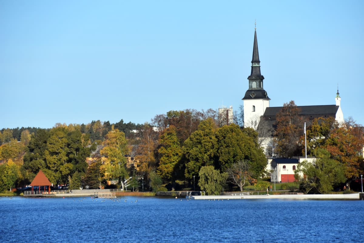 En bild tagen från vattnet som visar en bred vy över en strandlinje med höstträd och några byggnader under en klarblå himmel. Ett vitt kyrktorn med svart spira dominerar den högra sidan av horisonten.
