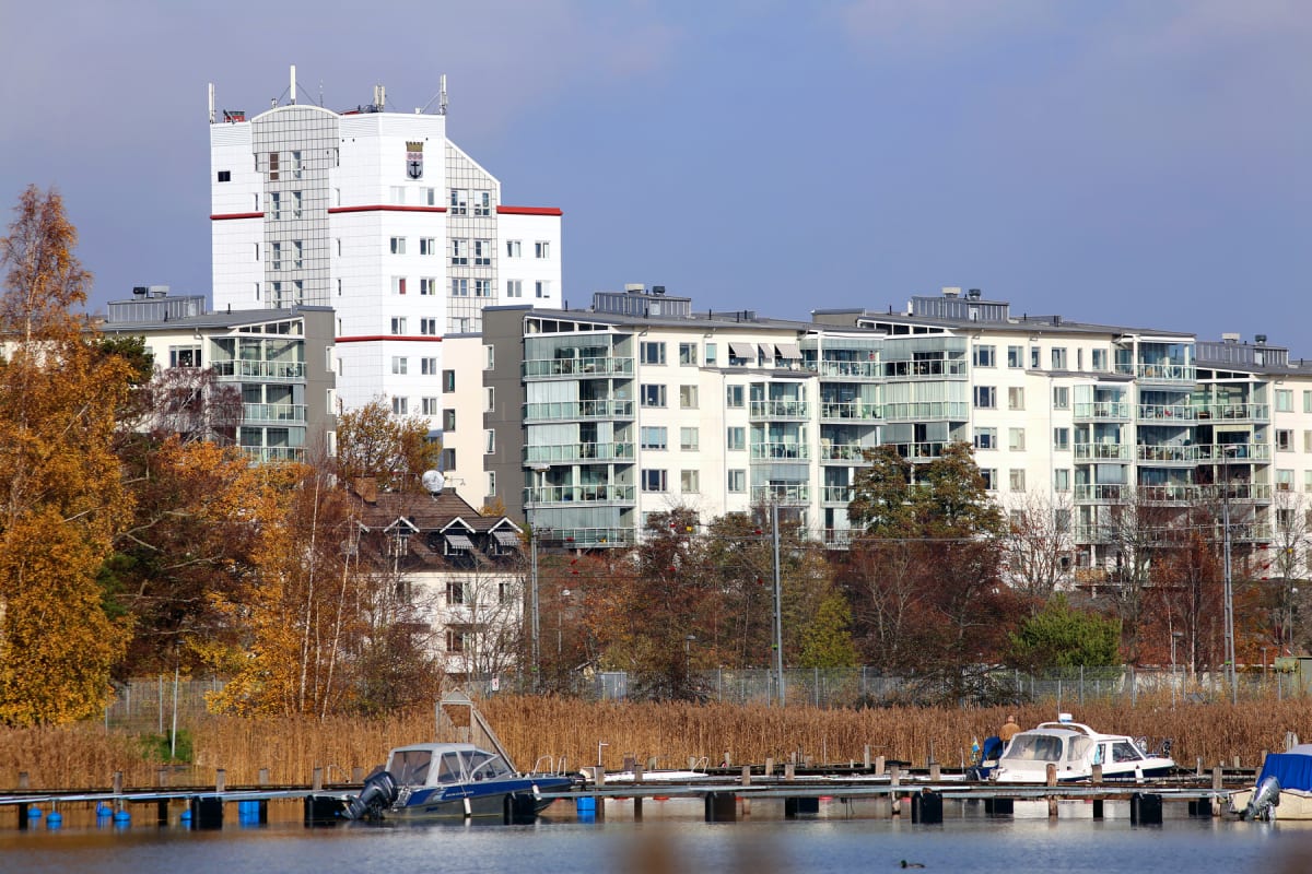 En bild som visar en småbåtshamn med två motorbåtar förtöjda vid en brygga i förgrunden, omgiven av vass. Bakom vassen står träd med höstfärger, och i bakgrunden syns flera flerbostadshus i olika storlekar och stilar under en klarblå himmel.