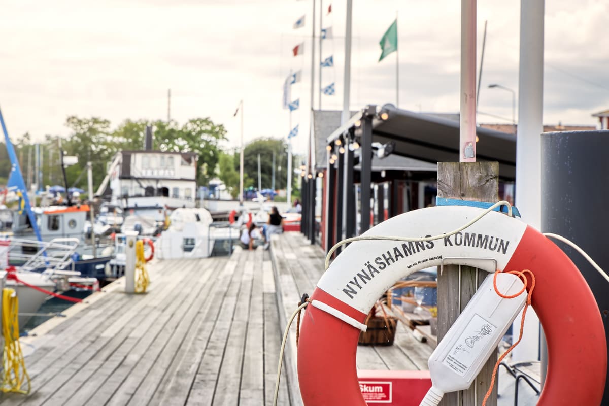En livboj med texten "Nynäshamns kommun" syns i förgrunden, fastsatt vid en trästolpe på en brygga. I bakgrunden syns båtar förtöjda längs bryggan, byggnader och människor. Flaggor syns på flaggstänger och himlen är molnig