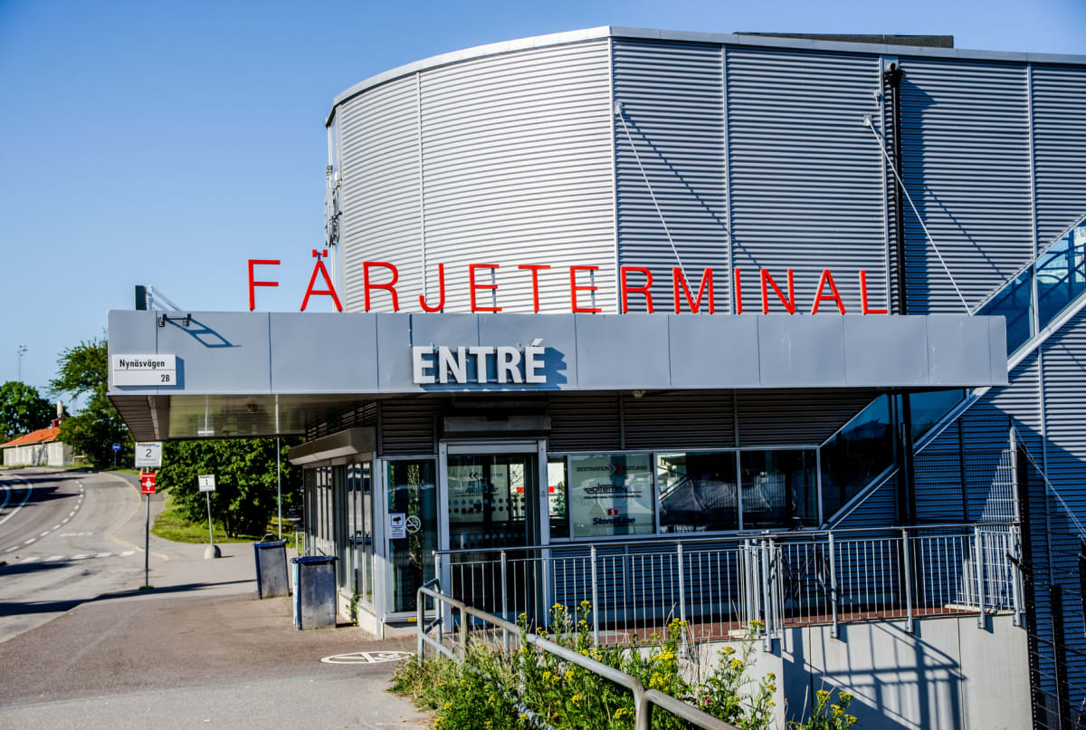 En modern byggnad med en “färjeterminal” skylt i röd text och en “entré” skylt i vit text syns under en blå himmel. Utanför byggnaden finns en asfalterad väg, en gräsmatta med vilda blommor och en vägskylt med texten Nynäsvägen 2B.