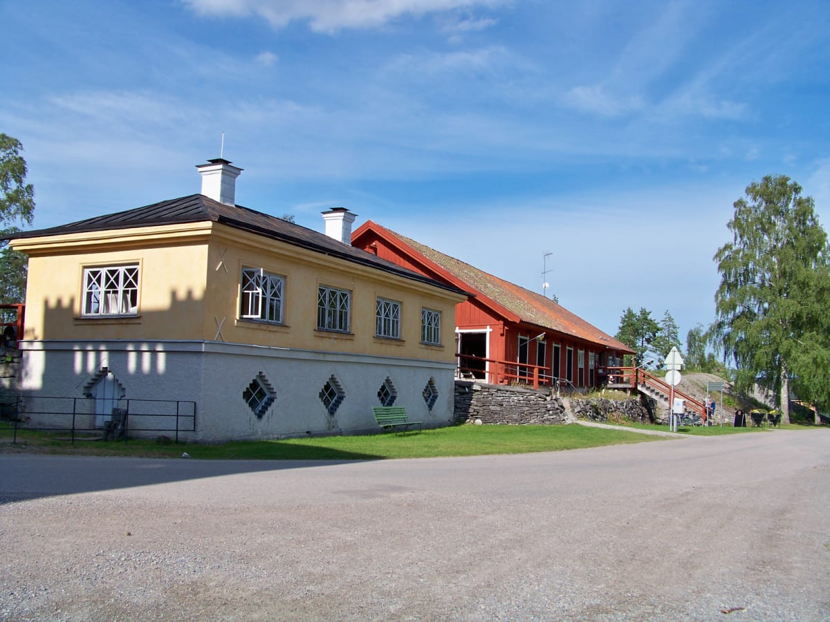 En bild som visar en gul byggnad med vita knutar och ett svart tak till vänster, och en röd byggnad med rött tak och en lång veranda till höger. Mellan byggnaderna syns gräsytor, en grusväg och en del av en asfalterad väg. I bakgrunden syns blå himme