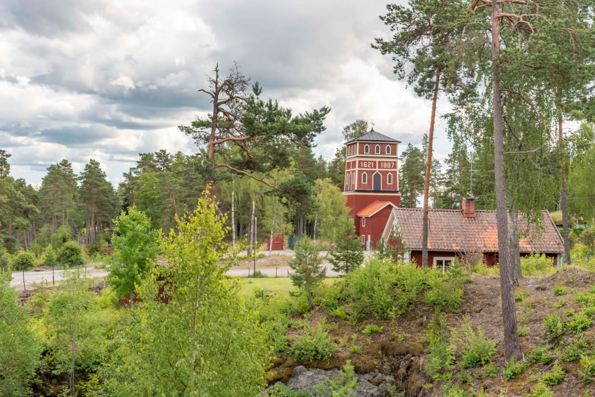 En bild på en röd byggnad med ett torn och siffrorna 1621 och 1887. Byggnaden omges av grönska, träd och buskar. I bakgrunden syns skog och en gråmulen himmel. Ett mindre rött hus med tegeltak finns bredvid tornet.