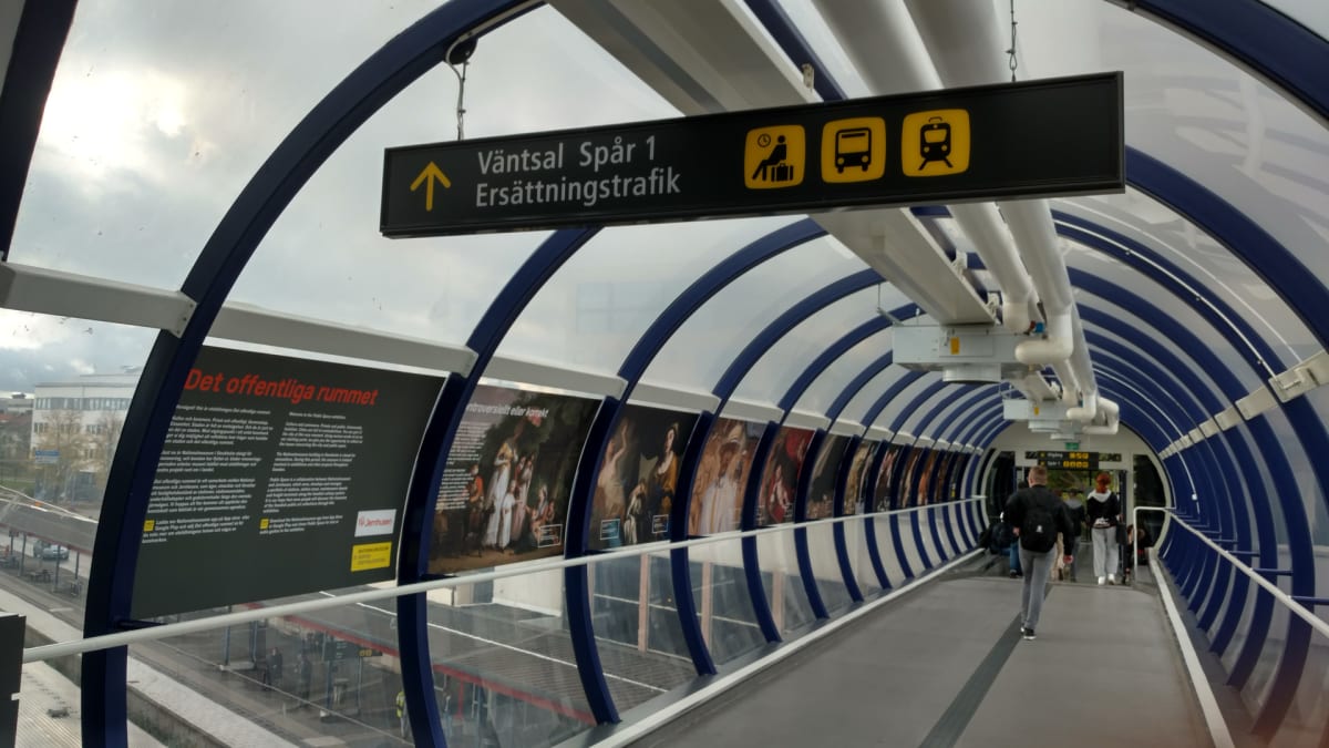Bilden visar insidan av en glaskorridor som fungerar som en gångbro, med blå bågar och genomskinliga väggar. En skylt indikerar "Väntsal Spår 1 Ersättningstrafik" och personer går längs korridoren. Utanför syns byggnader och tågspår.