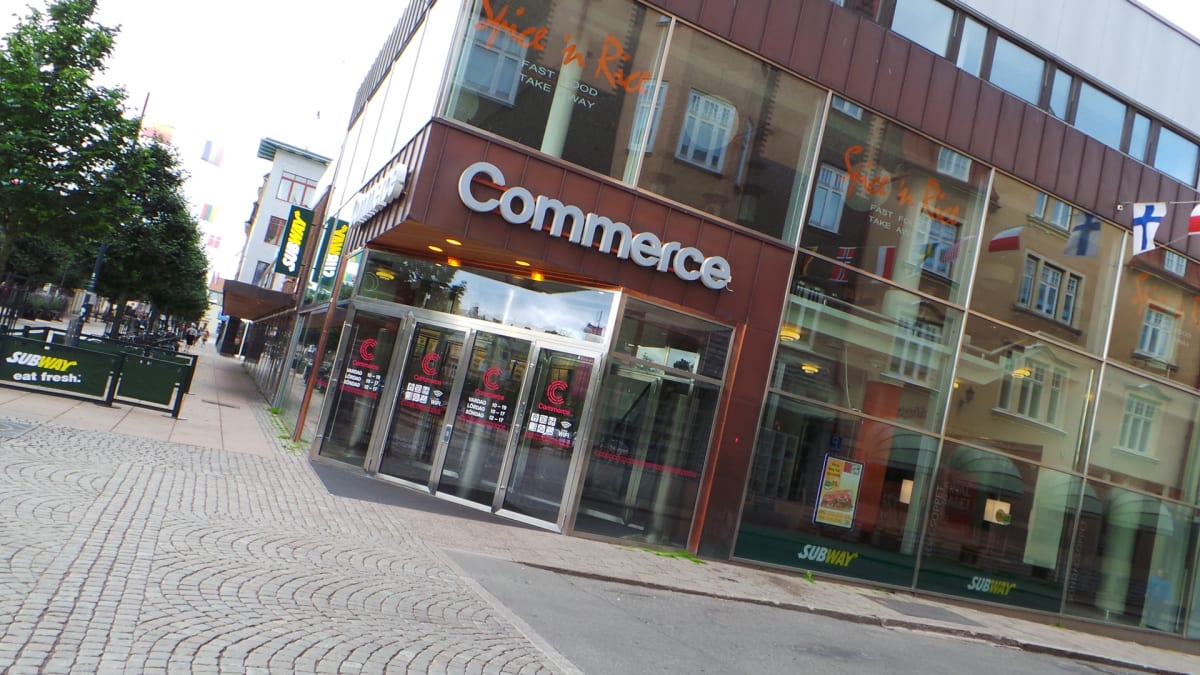 En bild som visar en byggnad med skylten "Commerce" och en Subway-restaurang på bottenvåningen. Fasaderna är gjorda av glas och reflekterar omgivande byggnader och himlen. En trottoar i sten leder förbi byggnaden, med träd längs kanten.