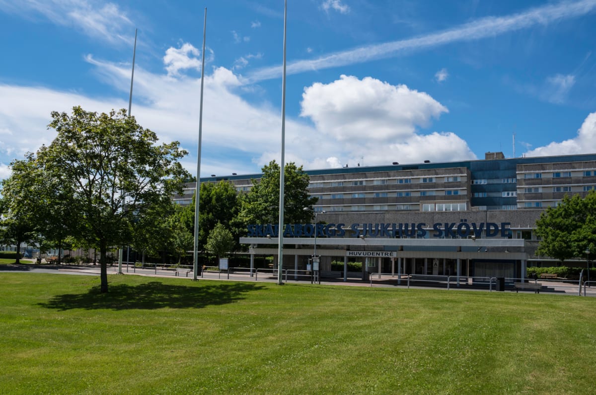 En bild tagen utomhus framför en stor sjukhusbyggnad i flera våningar med "Skaraborgs Sjukhus Skövde" skrivet på fasaden och "Huvudentré" ovanför ingången. En gräsmatta och flera träd syns i förgrunden, samt tre höga flaggstänger. Himlen är blå med v