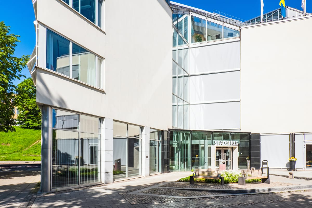 En modern byggnad med vita fasader och stora glaspartier under en klarblå himmel. Framför ingången, märkt "STADSHUS", finns bänkar, planteringar och en stenlagd yta. En svensk flagga syns på taket.