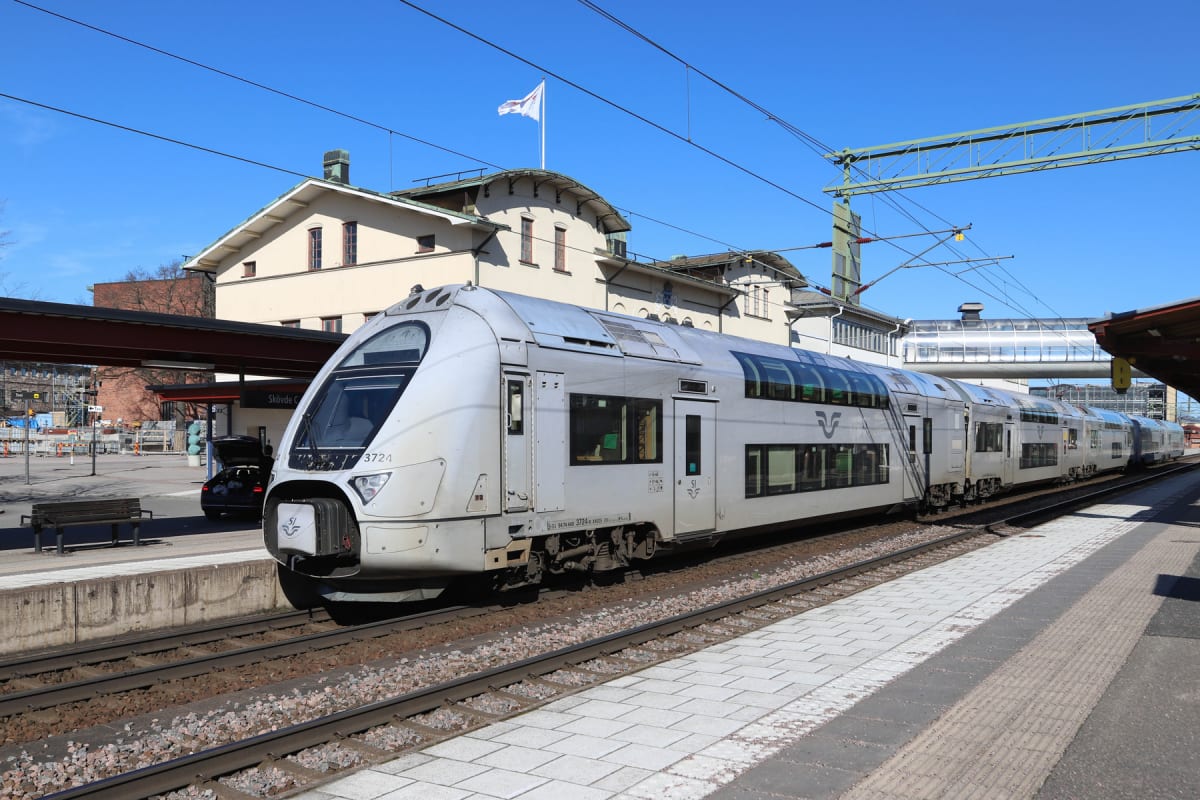 En silverfärgad dubbeldäckare från SJ står vid en perrong. I bakgrunden syns en ljus byggnad med flaggstång och en blå himmel. Järnvägsspår och en bil med öppen bagagelucka är också synliga.