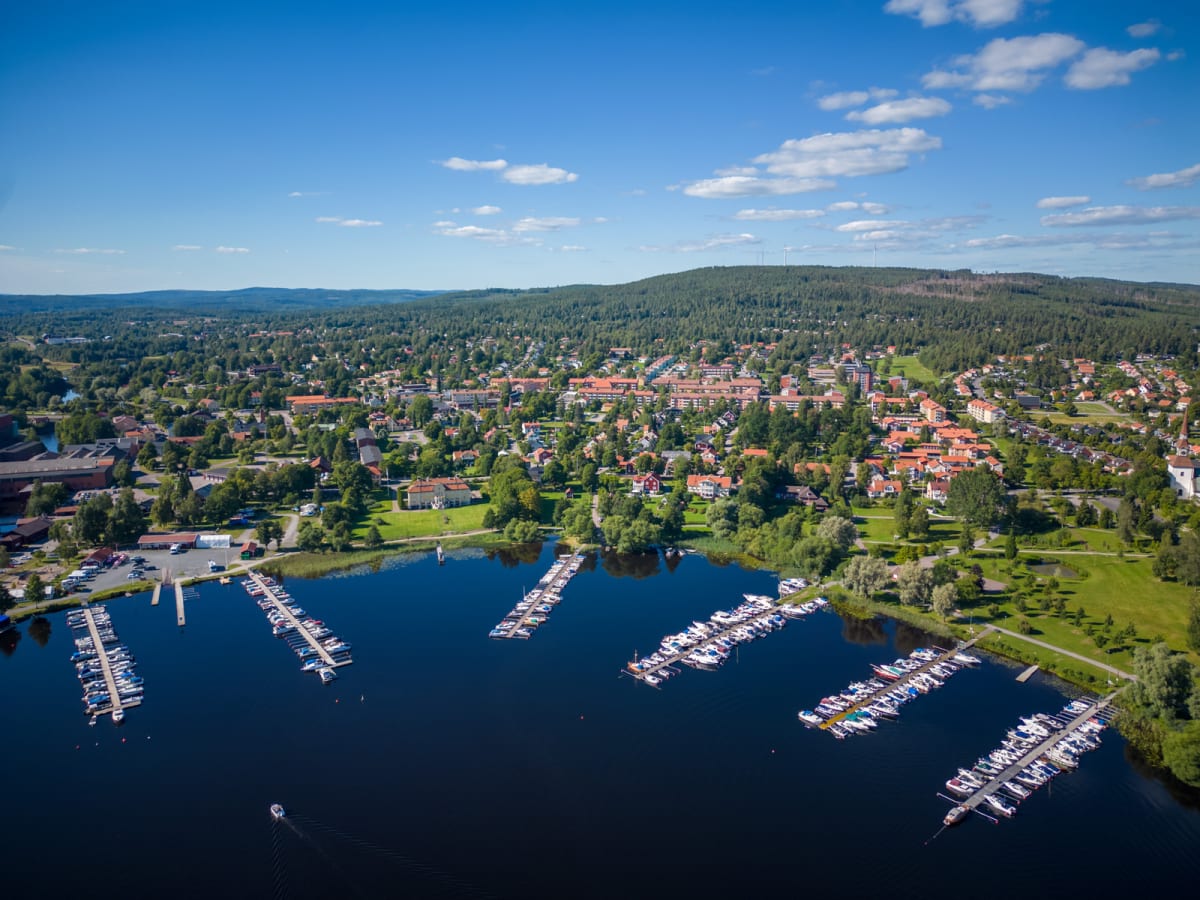 En vidvinkelbild tagen uppifrån över en sjö med ett flertal båtar förtöjda vid bryggor. Längs sjöns strandlinje syns en tätbebyggd stad med bostadshus, grönområden och skogklädda höjder i bakgrunden under en klarblå himmel med några moln.