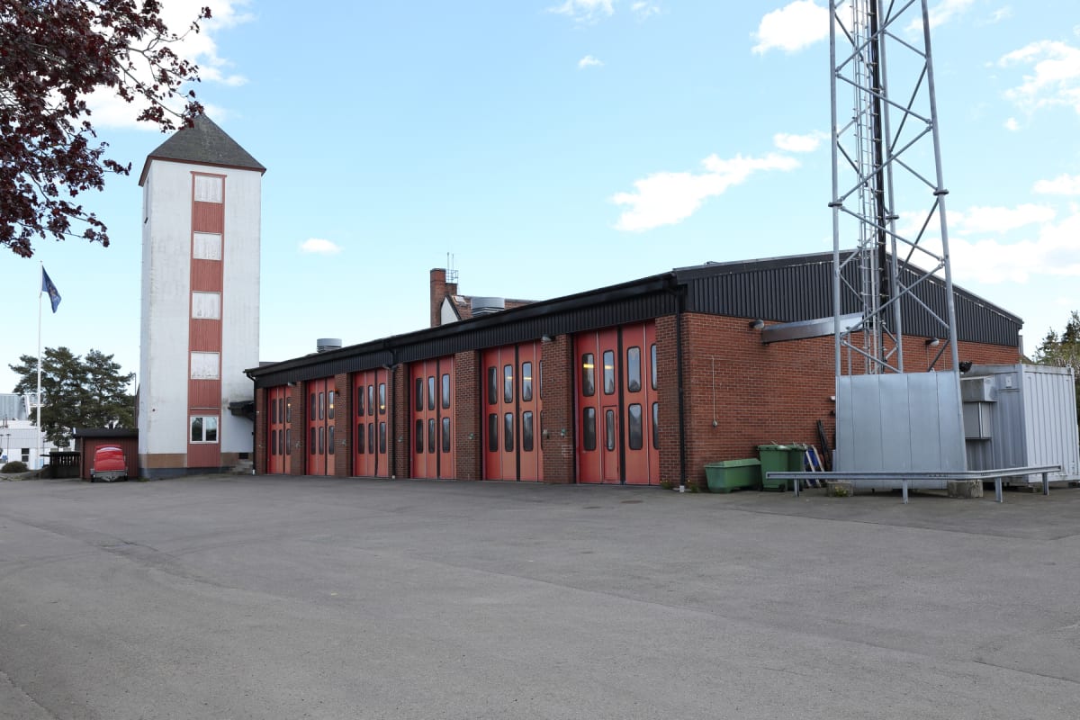 En bild som visar en brandstation med en vit byggnad och rött tak i förgrunden, bredvid en tegelbyggnad med flera röda portar. Till höger om tegelbyggnaden finns en metallmast, och på marken framför byggnaderna syns en asfalterad yta.