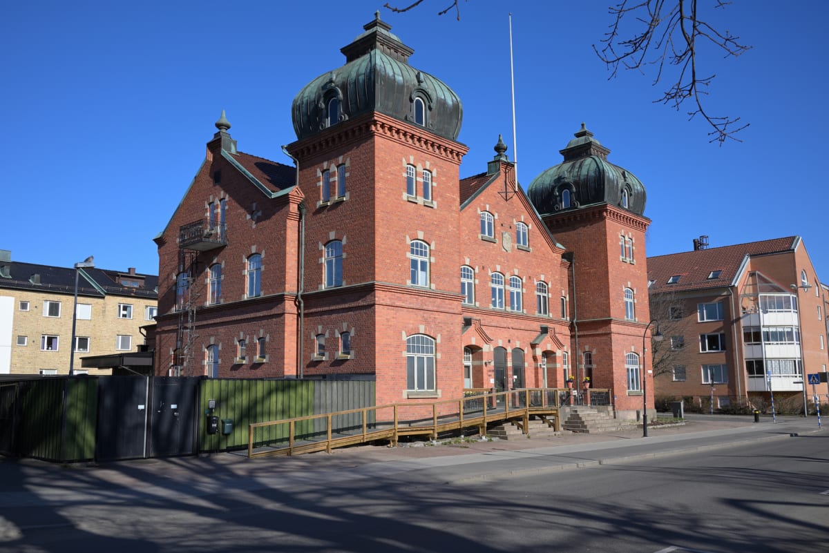 En stor, röd tegelbyggnad med mörka kupoltak och vita fönsterkarmar dominerar bilden. Byggnaden har en ramp som leder upp till entrén. På vänster sida syns en grön inhägnad, och i bakgrunden finns andra byggnader samt en klarblå himmel.