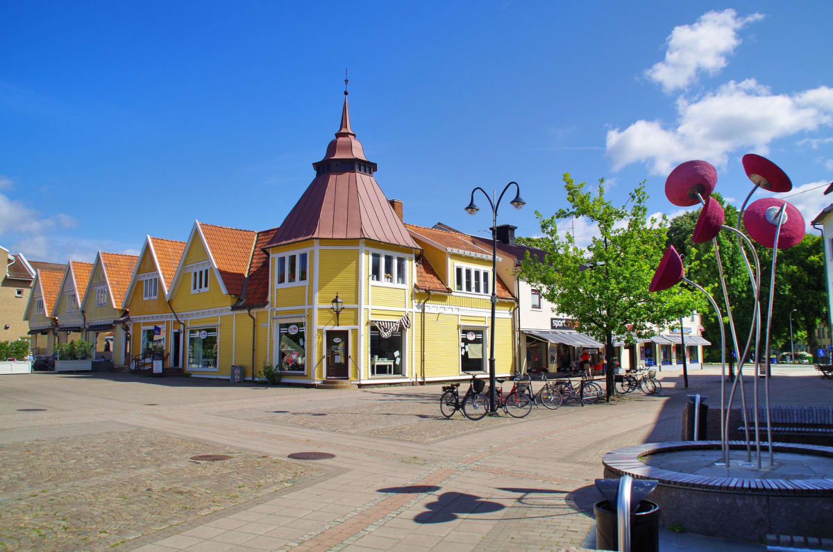 En ganska bred bild av en öppen plats framför en rad byggnader med gul fasad och orange takpannor. En av byggnaderna har ett runt torn med spetsigt tak. Flera cyklar är parkerade vid en lyktstolpe. Till höger syns en skulptur med runda, röda former.