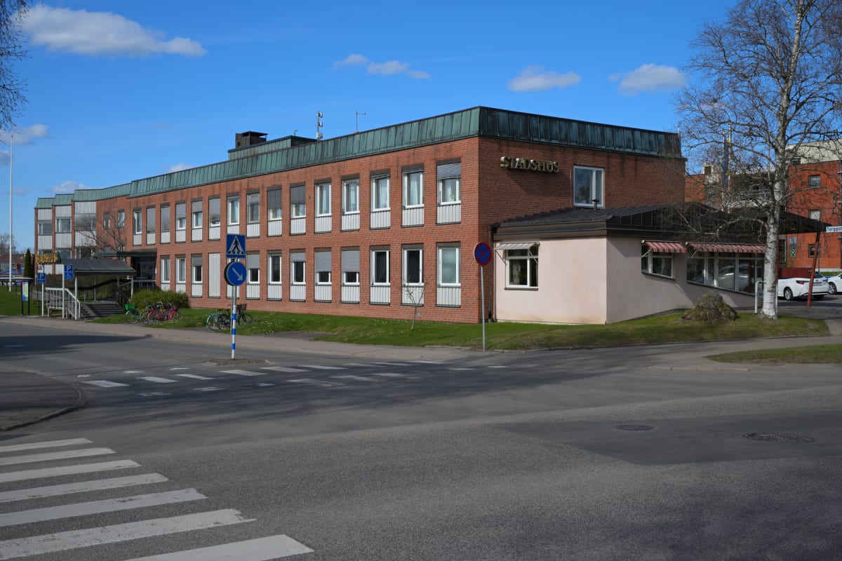 En stor röd tegelbyggnad med vita fönster och mörkgrönt tak, med texten "STADSHUS" på fasaden. Framför byggnaden finns en väg med övergångsställen, trafikskyltar, gräsytor och parkerade cyklar. Himmelen är klarblå med några vita moln.