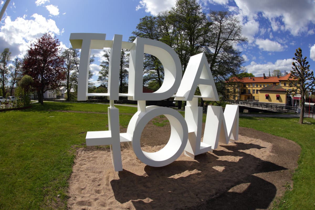 En stor vit skylt med texten "TIDAHOLM" står på en sandplätt, omgiven av en gräsyta med träd i bakgrunden. Ett gult hus med rött tak och en bro syns i fjärran under en blå himmel med moln.