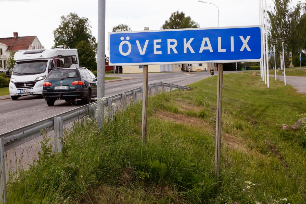 En blå skylt med vit text "ÖVERKALIX" står vid en vägkant. På vägen kör en vit husbil och en mörkgrön kombi. I bakgrunden syns byggnader och träd under en molnig himmel. Gräs och vildblommor växer i förgrunden.