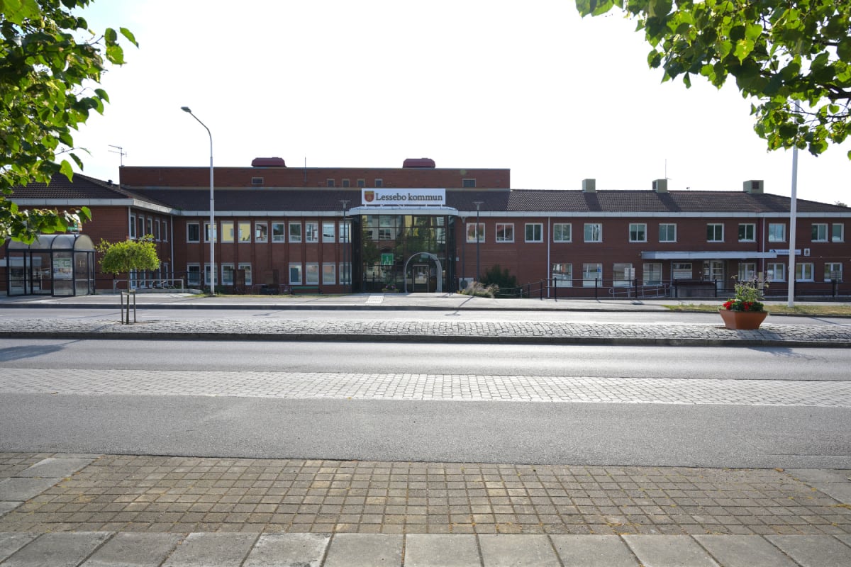 En fasadbild av kommunhuset i Lessebo kommun, sett över en gata med en busshållplats och ett blomsterarrangemang. Byggnaden är huvudsakligen i rött tegel med en glasentré i mitten och grönska syns i förgrunden och sidorna av bilden.