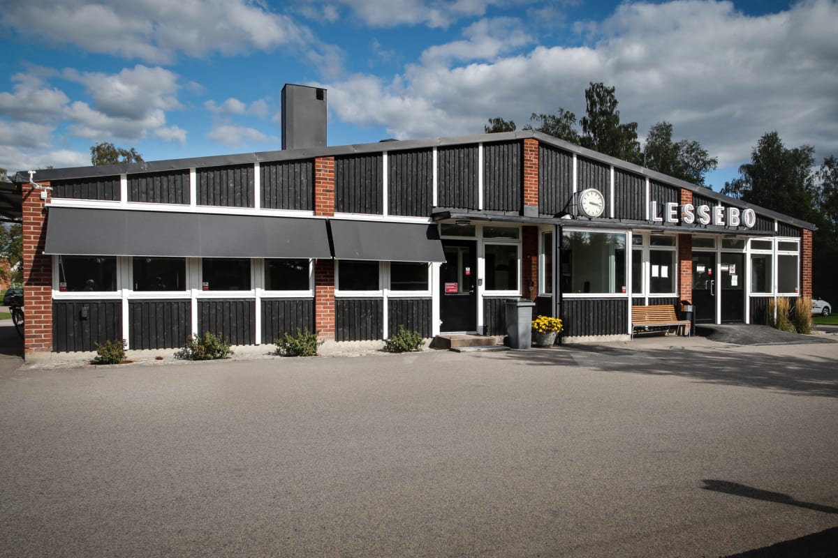 En enplansbyggnad med mörk träpanel och tegelpartier, vita fönsterkarmar och svarta markiser. På byggnaden syns texten "LESSEBO" och en klocka. Utanför finns en asfalterad yta, buskar och blomkrukor.