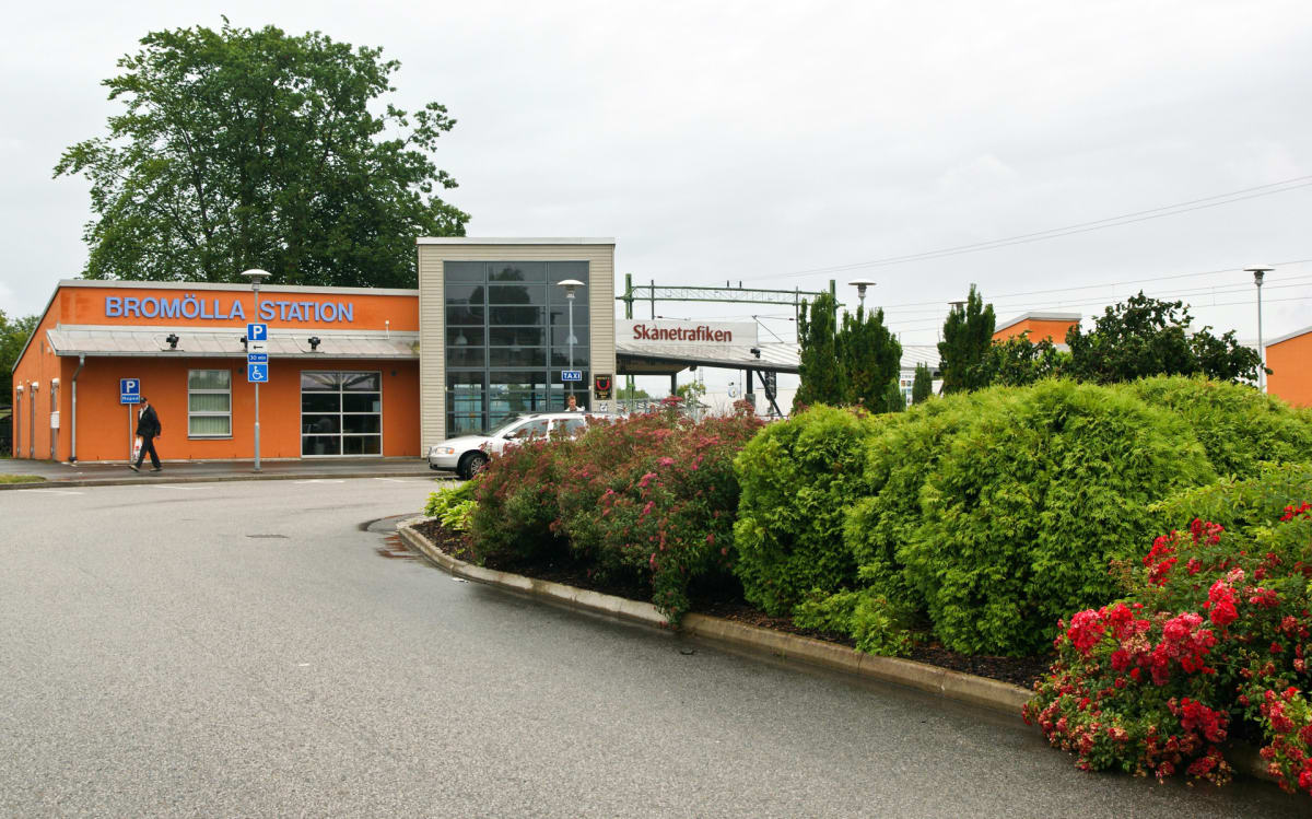 En bild av Bromölla Station med en orange byggnad med blå text och en glasfasad. En person går på trottoaren och en vit bil står parkerad. Växtlighet, inklusive buskar med röda blommor, syns i förgrunden och intill en väg. Himlen är mulen.