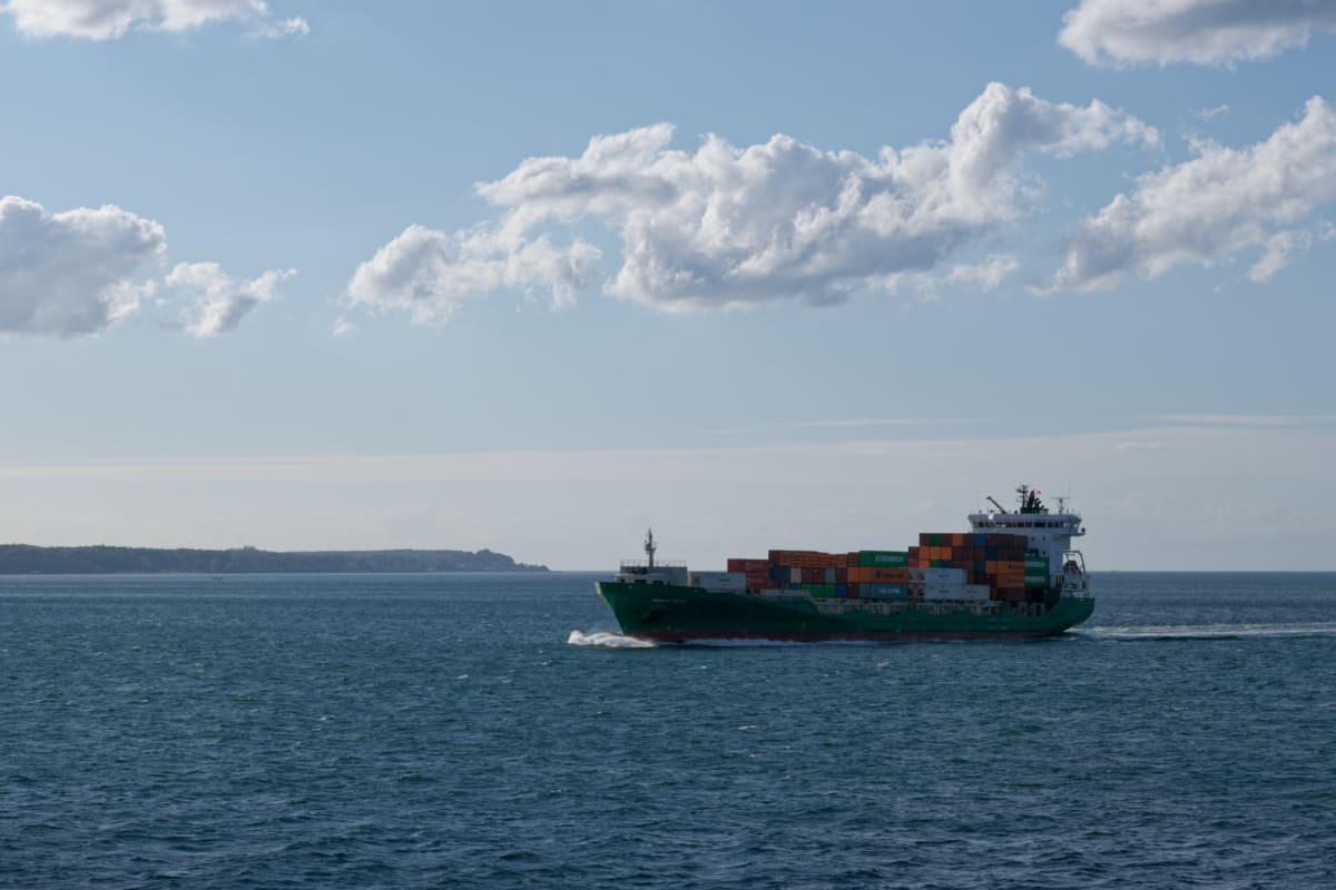 En stor grön containerbåt med flera containrar i olika färger färdas på ett blått hav under en klarblå himmel med spridda vita moln. I bakgrunden syns en grön landmassa under horisonten.
