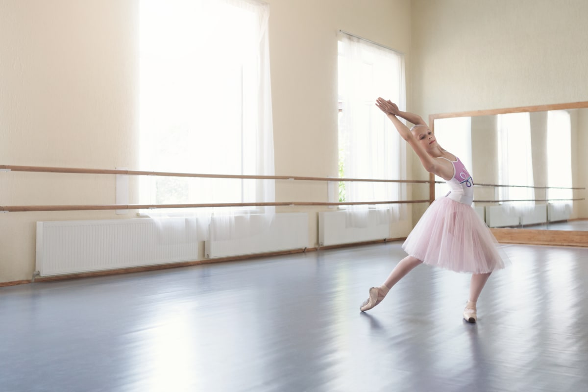 En ung ballerina utför en pose i en dansstudio. Hon bär en vit och lila dräkt med en rosa tyllkjol och tåskor. I bakgrunden syns stora fönster med vita gardiner, element och en spegelvägg med träramar.