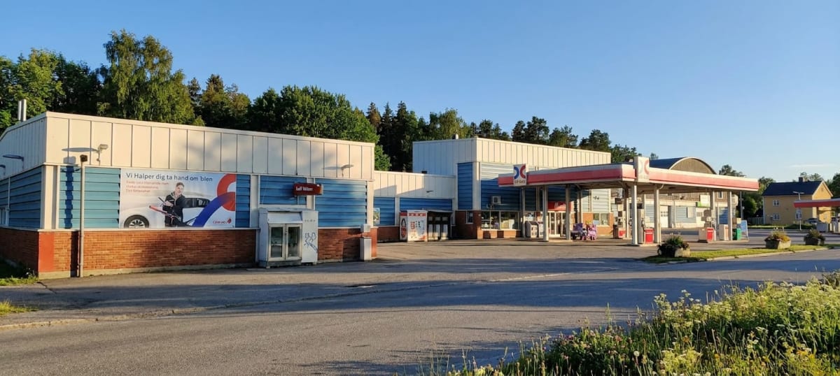 En bild över en bensinstation med en byggnad som har en reklamationsskylt med texten "Vi Hjälper dig ta hand om bilen" och en man som håller i en spak. Stationen har tak över bensinpumparna. Flera byggnader och träd syns i bakgrunden under en klarblå
