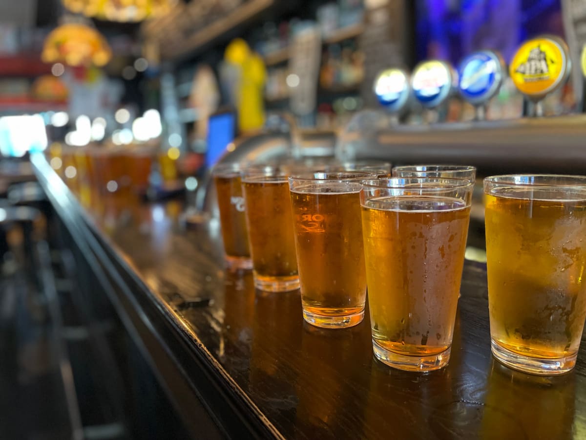 En rad med sju glas fyllda med en gulbrun vätska står på en mörk, blank träbänk. Glasen visar kondens på utsidan. I bakgrunden syns suddiga detaljer av en bar, inklusive några ölkranar med runda logotyper.