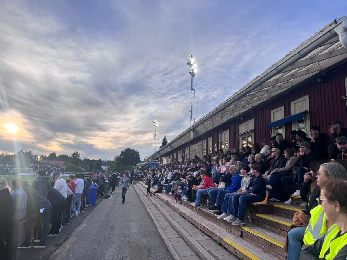 En utomhusbild som visar en fullsatt läktare med publik på en sportanläggning under en kväll. Flera personer sitter på träläktare med tak, medan andra står längs ett staket. Strålkastare är tända och solen är på väg ner i horisonten.
