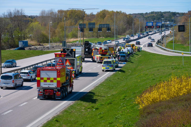 Bilden visar en större väg, sannolikt en motorväg eller större landsväg, där trafikolyckor har inträffat. Flera utryckningsfordon, inklusive brandbilar, polisbilar, ambulanser och underhållsfordon med varningsskyltar, står parkerade på den högra körb
