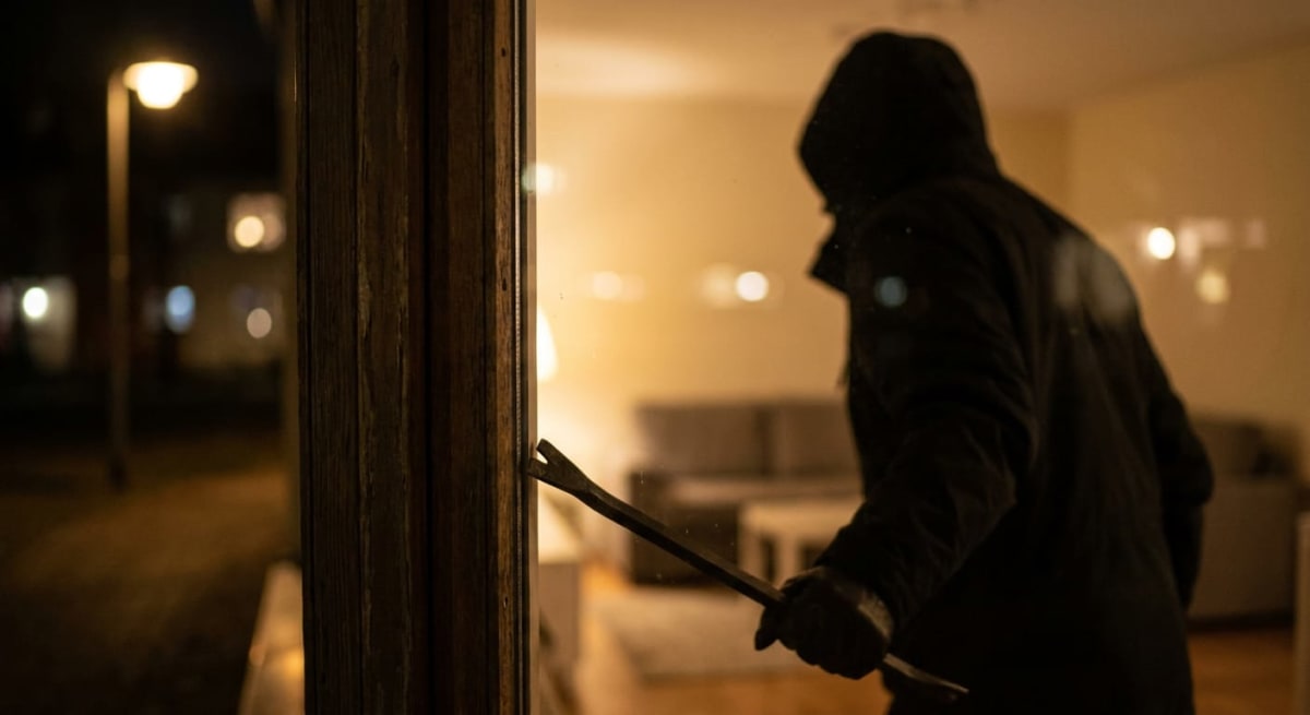 En person klädd i en mörk huvtröja och handskar håller ett kofotsliknande verktyg mot en dörr- eller fönsterkarm. Verandans glas reflekterar personens silhuett. Bakgrunden utanför är mörk med suddiga ljuspunkter från gatlyktor, medan ett upplyst rum
