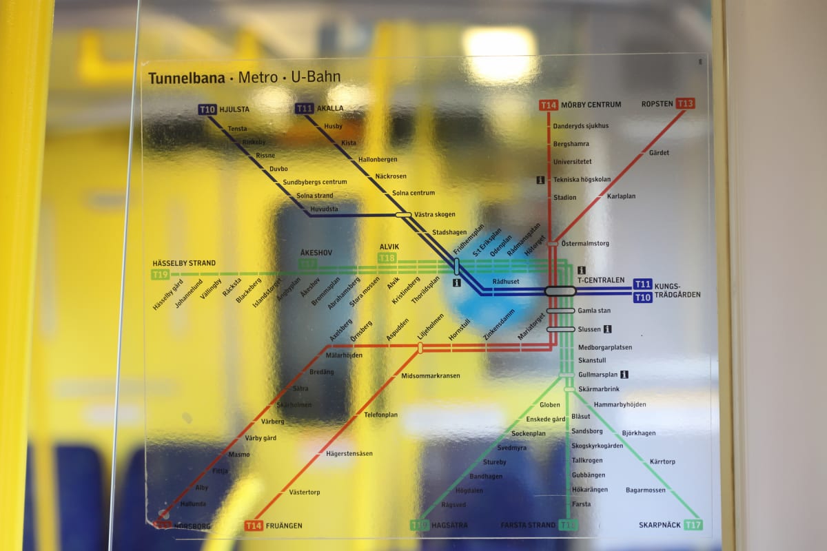 En tunnelbanekarta över Stockholms tunnelbana, fäst bakom ett transparent skydd, visar linjer och stationer med texten "Tunnelbana • Metro • U-Bahn" högst upp. Kartan reflekterar starka gula ljus, troligtvis från insidan av ett tunnelbanetåg.