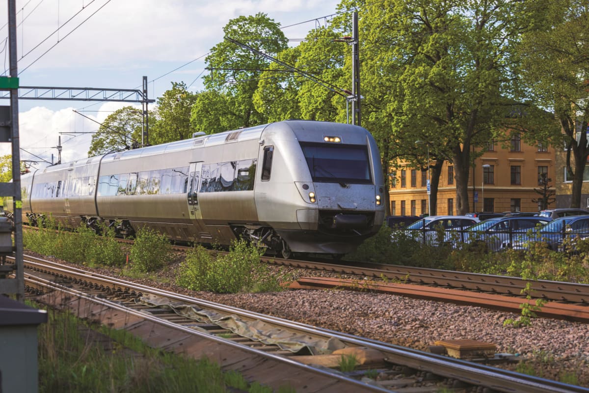En silverfärgad persontåg syns på spår, med gröna träd och en gul byggnad i bakgrunden. Ett flertal bilar står parkerade bakom ett staket vid den gula byggnaden. Blå himmel med moln syns ovanför trädkronorna.